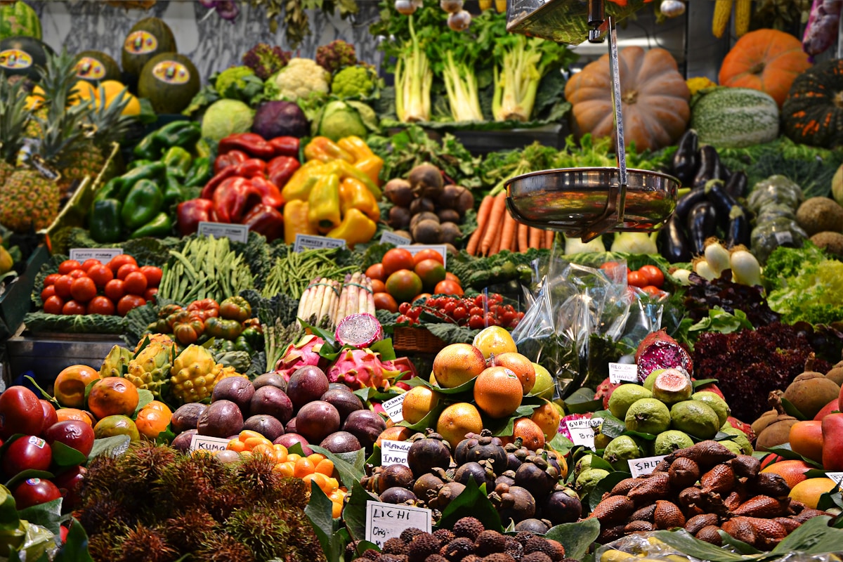 Market stall piled with mangosteens, dragon fruit, rambutan, pumpkins, bell peppers, and carrots beneath a hanging metal scale